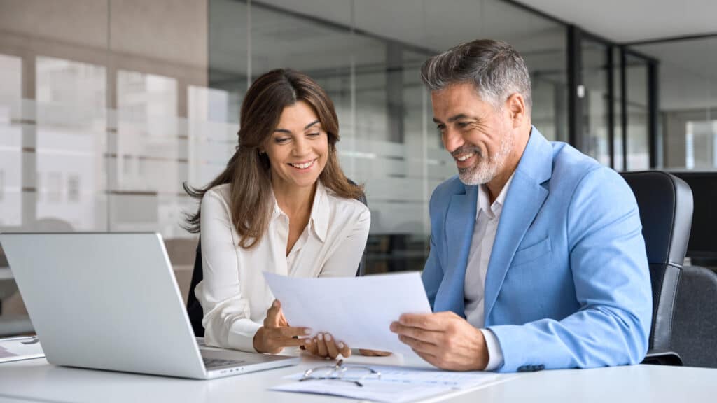 Two business professionals reviewing documents together at a modern office desk with a laptop and papers.